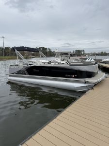 Pontoon boats docked at a wooden pier on a calm body of water under an overcast sky, with trees in the background of a marina.